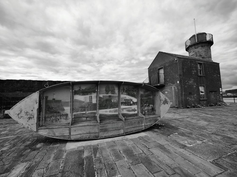 A black and white view at a stone harbour. There is a wooden boat on its side and a stone building to the right of it.
The boat is "Visions of a Harbour Past," a public art installation by Stephen Broadbent located at Whitehaven Harbour in Cumbria, England. 
The artwork features a boat-like structure with glass panels containing historical images of Whitehaven's past, particularly focusing on the town's maritime heritage and the local Rocket Brigade.
The building to the right of the boat is the former headquarters of the town's Rocket Brigade.
Whitehaven Volunteer Rocket Brigade was formed in 1849. The volunteers would haul their equipment to the coast where a ship was in distress. Rockets attached to a line were fired over the vessel, and then a much heavier cable could be drawn across, allowing the use of a Breeches buoy system to rescue passengers and crew.