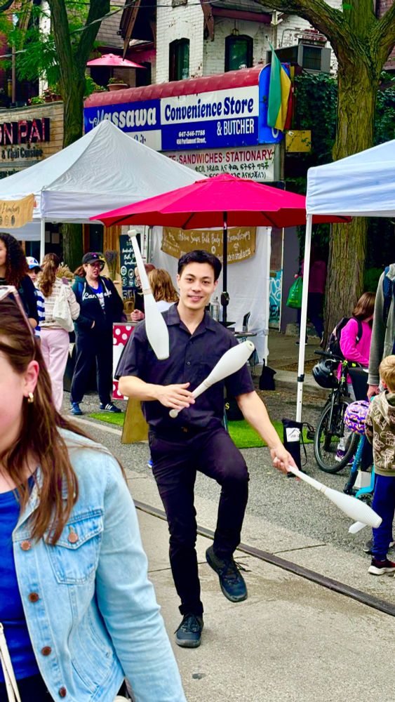 A man wearing black juggling white pins on the street while wearing a huge smile