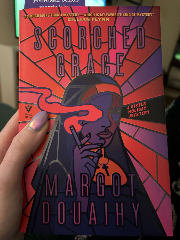 The cover of the fiction book ‘Scorched Grace’ by Margot Douaihy. The cover features a nun who is wearing a cross around her neck, with a cigarette in her right hand, and a stern/moody face. The colour palette is pink, purple and indigo, with blocks of sun rays extending out from the middle as the background. The book cover also says ‘A Sister Holiday mystery’.
