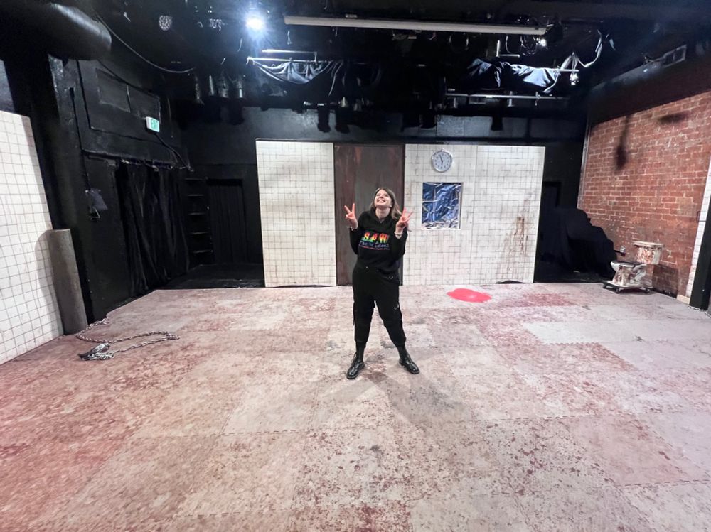 A stage, with white tile flats, a very dirty and bloody floor, and a nasty toilet on the right. A woman, holding up two peace signs, very excited to be there bc she finished up the first show running backstage. 