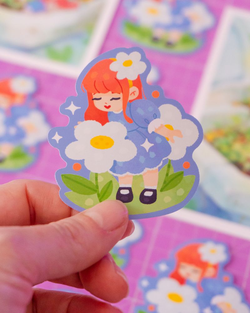 a hand holding a sticker of a cute red-haired girl sniffing a giant daisy flower