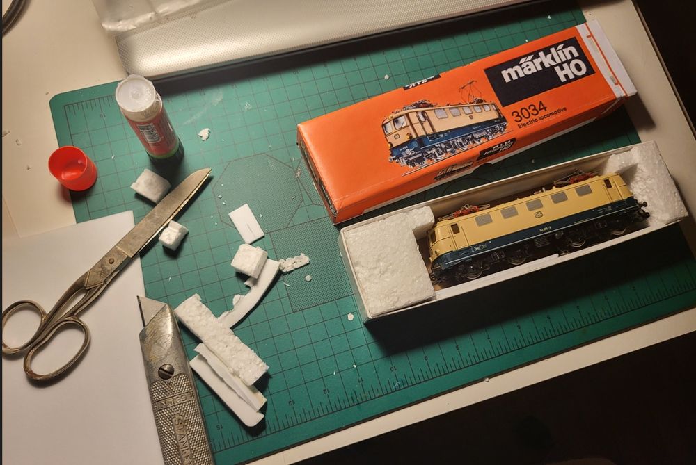 A Marklin 3034 in a box with an orange cover, on a table with glue, Scissors and Knife nearby. 