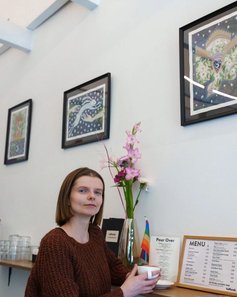 A woman with short brown hair sitting at a cafe table, holding a cup, with framed artwork on the wall behind and a colorful flower arrangement.