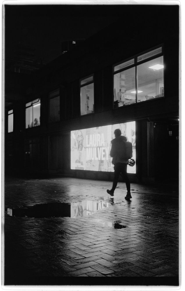 a person walking, silhouetted against a brightly glowing video billboard advertising a laura mulvey season at the bfi. it's also reflected in a puddle on the ground