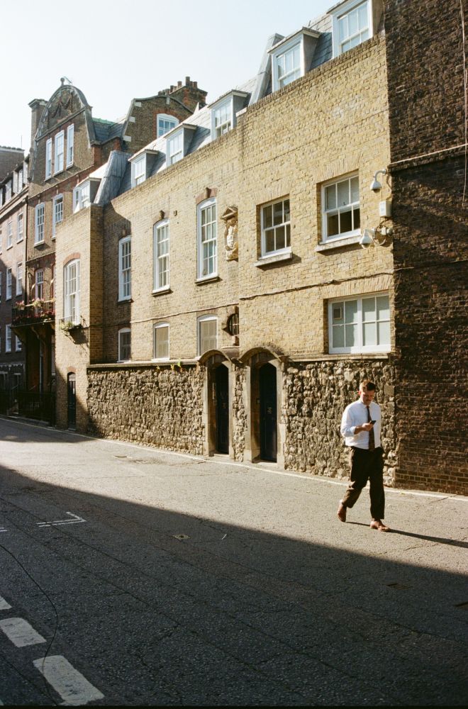 colour film photo, late afternoon, central london street. a man in a tie walks to the right, casting a long shadow, checking his phone. buildings are old brick, some sandstone and rough stone walls. two small doors near each other centre frame