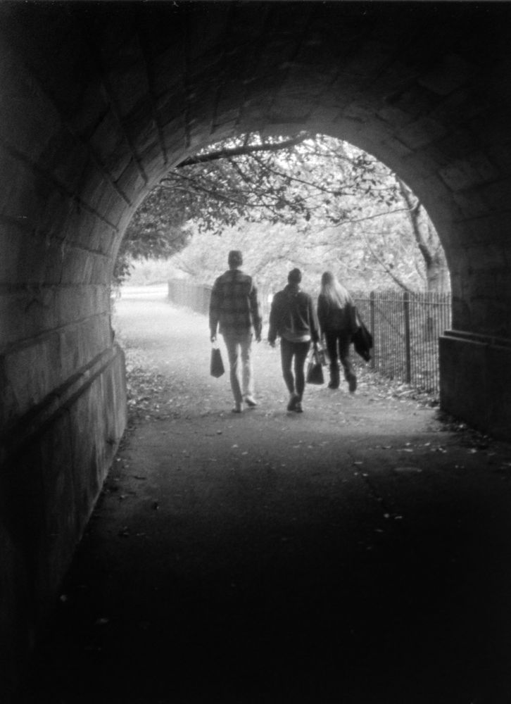 view from inside darkened tunnel out through circular mouth into urban parkland. three people, all carrying bags, are walking away just past the exit