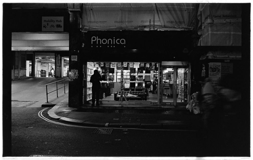 record store in soho at night. blurred bike rider to right. entrance to parking area off to left. a person standing in front of the window