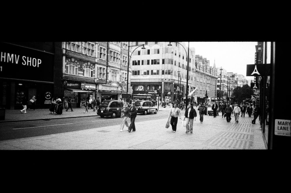 black and white film panorama of central london street scene. to the far left across the street is the hmv shop. black cabs. bunch of people walking with shopping bags. jd sports sign. far right half a road sign for marylebone lane