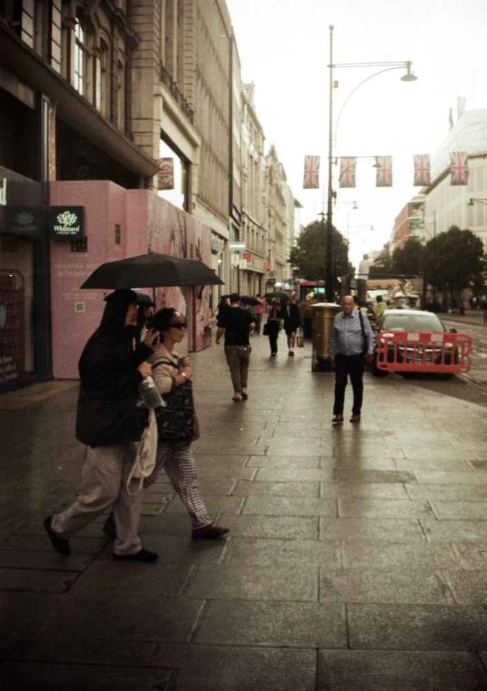 half frame film photo looking down rainy london street, two people with umbrella walk left to right