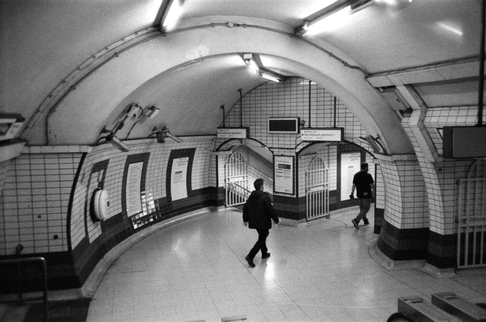 black and white film photo of arched passage in london underground station. it splits in two, and there are two figures heading right