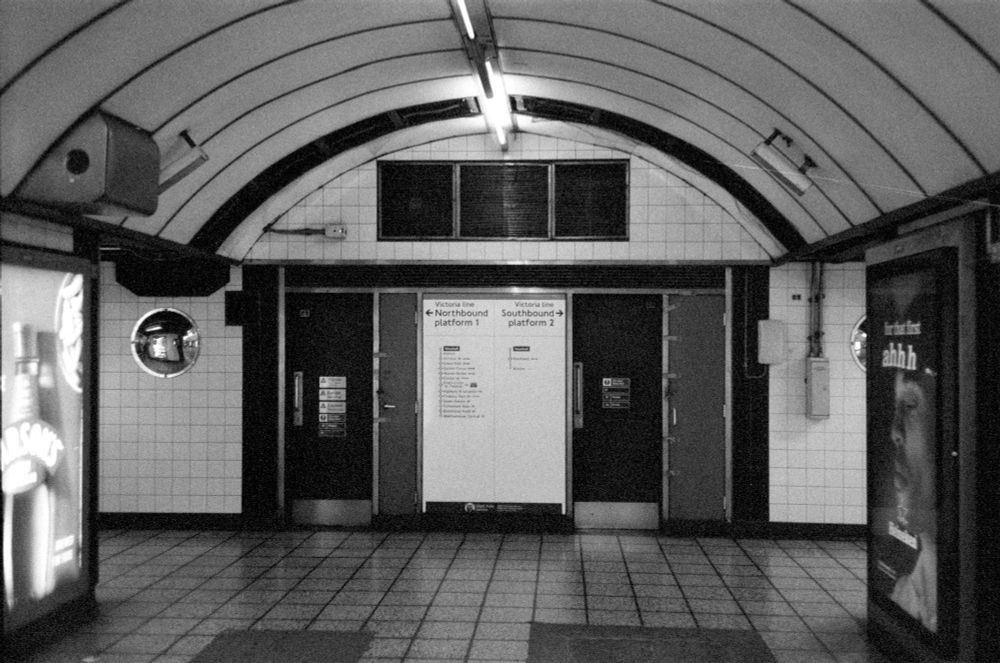 black and white film photo in an underground station at vauxhall. facing the end of a passage, where signs direct passengers left for northbound trains and right for southbound trains