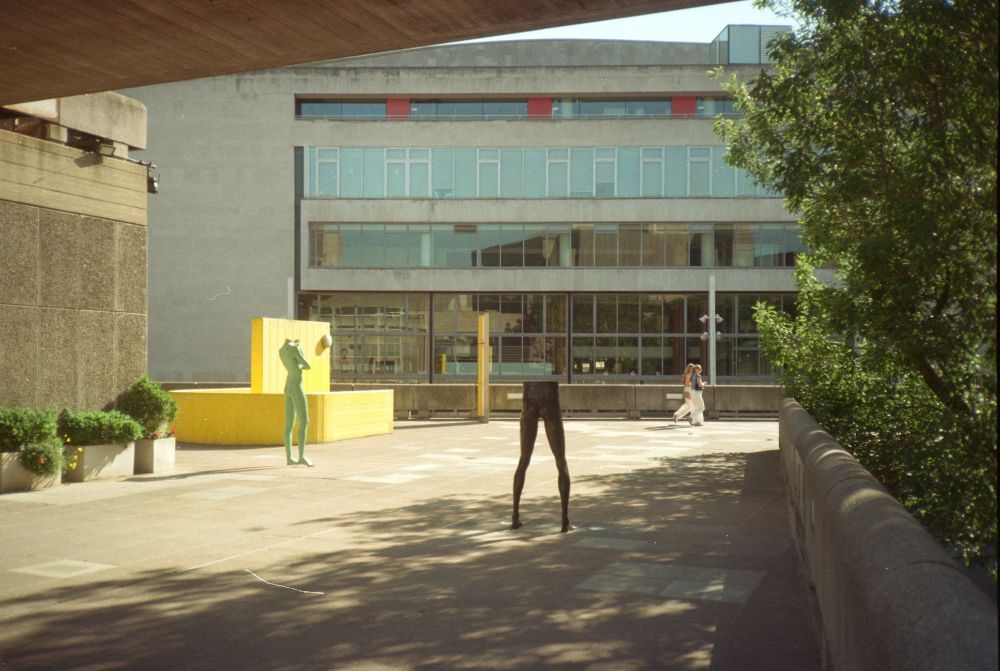 colour film photo outside hayward gallery, london. two incomplete sculptures, one just a pair of legs, the other missing a head, face away towards two people walking past in the distance. above and to the left is concrete, to the right a tree. bright yellow staircase in middle distance, furthest away is the royal festival hall