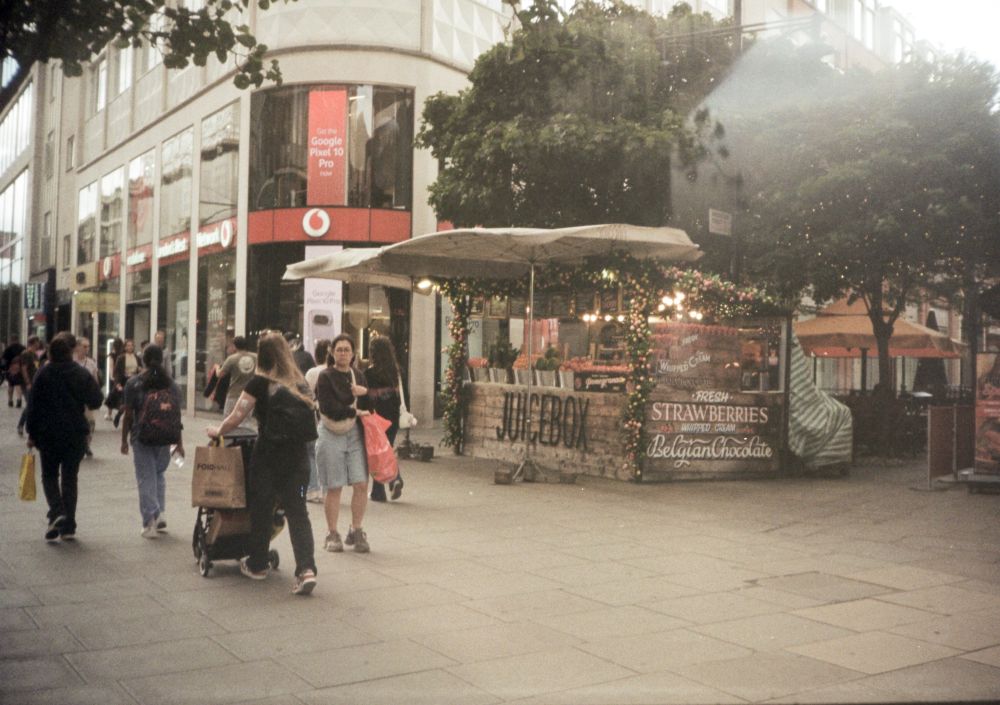 half frame film photo of london street with many people walking, a stall selling juice and strawberries with chocolate. some sort of double exposure appearing int he top right