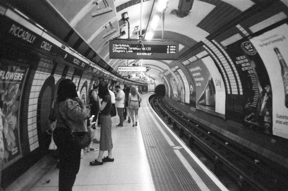 black and white film photo of london underground station platform. various people wait for a train. adverts line the right wall across the tracks. overhead is an information board, text strangely distorted