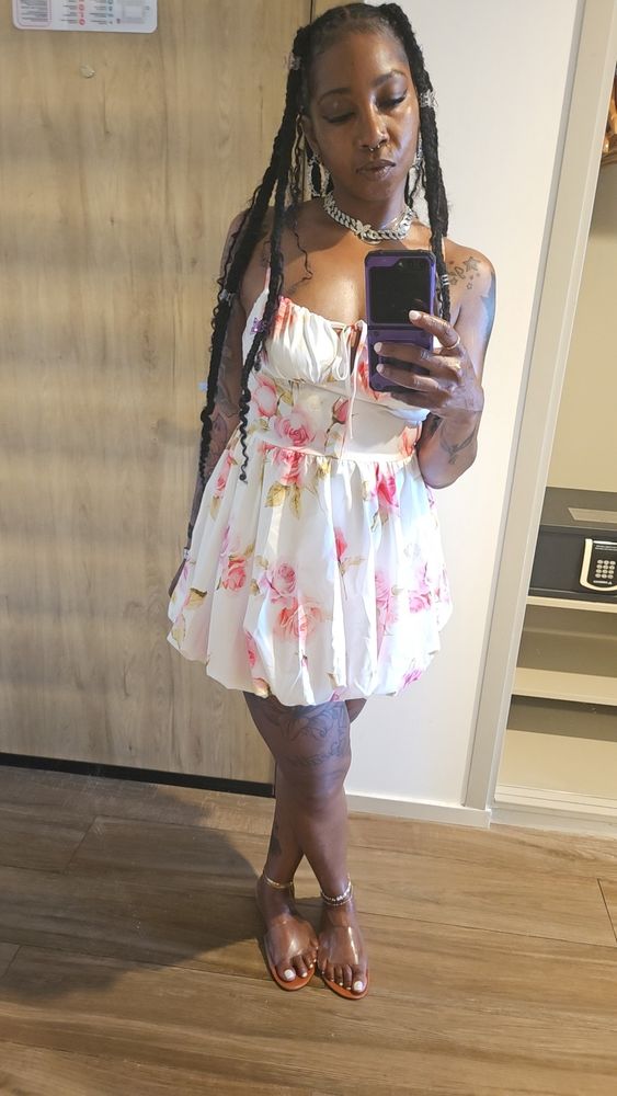 Super fine black woman wearing white dress with pink and green flowers taking a selfie. 
