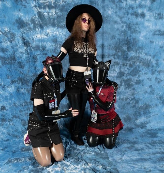 Two drones kneeling before their admin, One in a black dress, the other in red, clad in latex leggings and gloves. Both are wearing the same model gas mask, the Survivair Opti-fit. Their admin stands imposingly between them, wearing all black: short shorts, crop, latex leggings and gloves, a round rim hat and glasses. These drones love their admin very much.