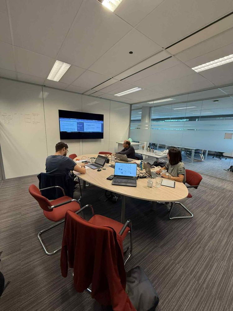 Three researchers work at a table with laptops, a large screen, in a bright room with glass windows