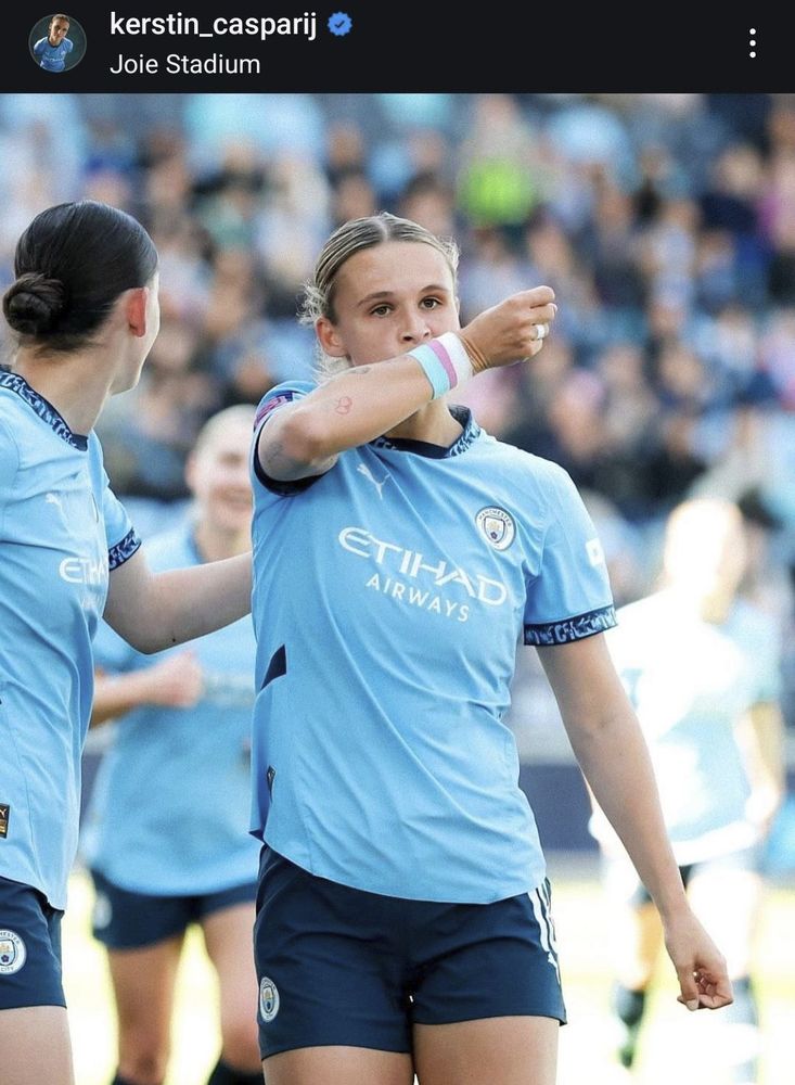 Kerstin Casparij im hellblauen Trikot Manchester Citys und am Handgelenk mit einem Armband in den Farben der Transpride-Flagge. Sie hält das Handgelenk mit dem Armband vor ihren Mund und schaut ernst zum rechten Bildrand. Neben und hinter ihr sind ihre Mitspielerinnen beim Torjubel zu sehen.