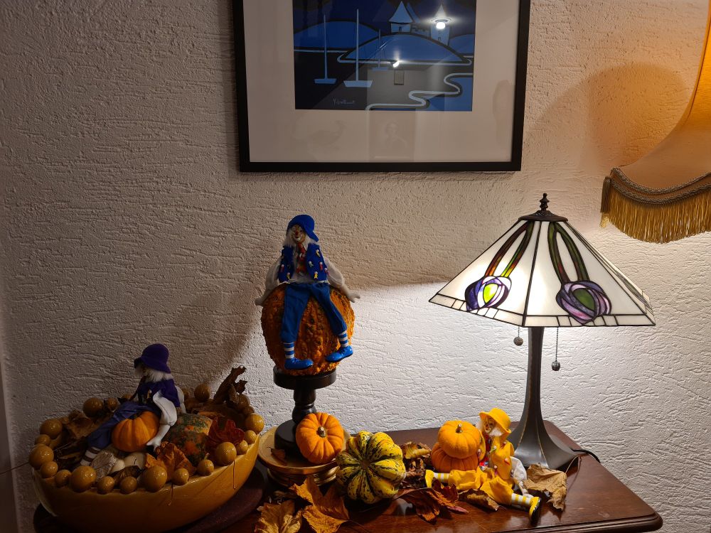 Three clowns sitting on various pumpkins surrounded by autumn leaves. Pumpkins are different shapes, sizes and colours.