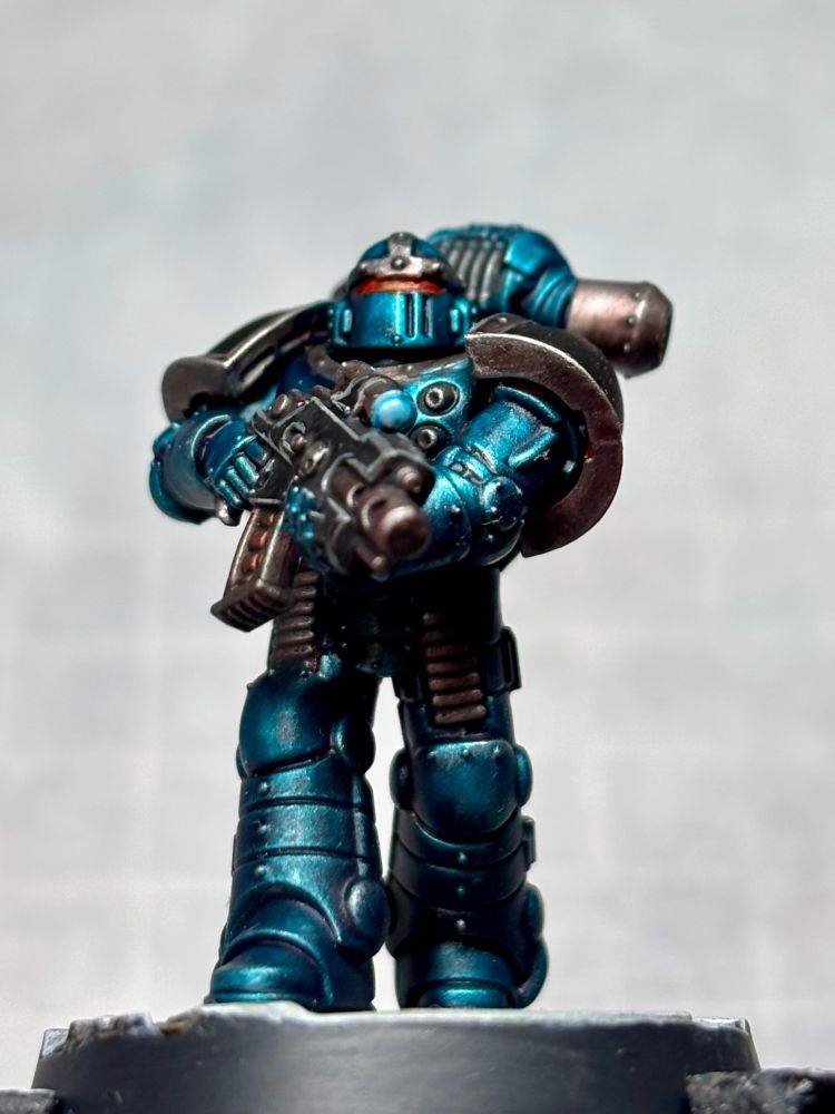 Space Marine legionary in Mk2 Crusade armour. In Alpha Legion colours, main colour is a metallic blue/green, shoulder pads in black with a steel metallic trim with a slightly warm tone. Eye lens is reddish orange in colour.