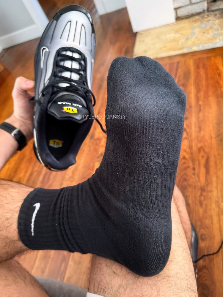 Sweaty, black Nike socks still on Tyler’s left foot, resting soles-up on his right knee, while he’s holding the black Nike Air Max Plus 3s he just pulled off his foot in his left hand.