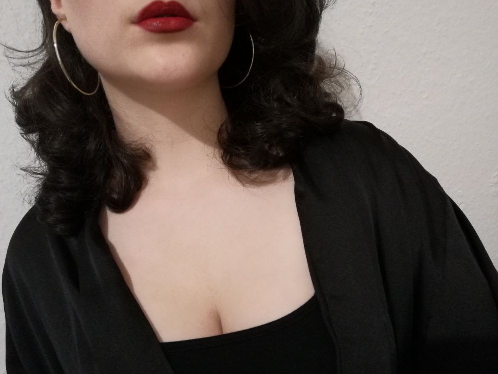 Selfie auf dem ich nur von meinen Lippen bis zu meinem Ausschnitt zu sehen bin. Ich trage roten Lippenstift, ein schwarzes Kleid, goldenen creolen und einen schwarzen satinumhang.