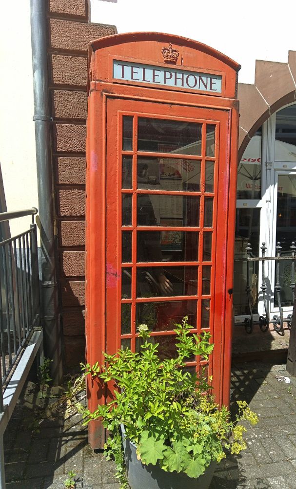 Rote Telefonzelle aus England 
Fenster unterteilt mit rechteckigen Streben 
Weiße Tafel oben an der Telefonzelle Schriftzug schwarz Telephone 
Davor eine grüne Pflanze 
Steht an einer Hauswand weiß mit braun abgesetzten Kacheln 