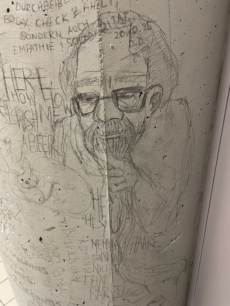 Eine Säule mit Kritzeleien. Man sieht einen Mann mit Brille der seinen langen Bart hält 