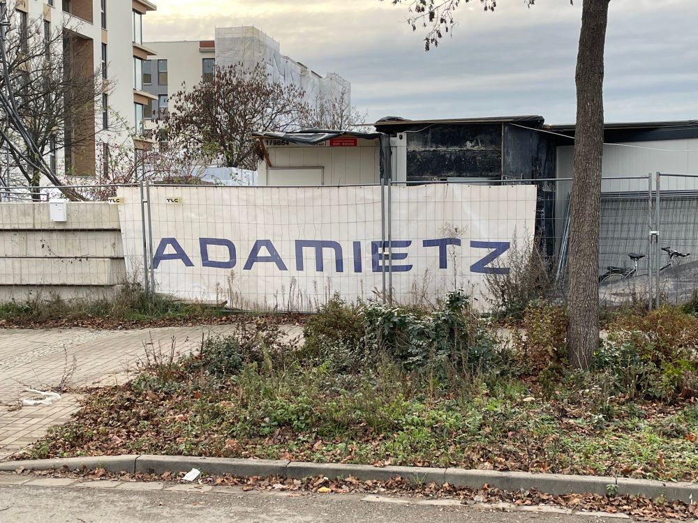 Ein Bauzaun an dem ein Banner einer Firma hängt. Darauf steht „Adamietz“
Daraus ergibt sich der Witz, dass es wie „Ah, da! Miez!“ klingt und somit wie ein Ausruf beim Anblick einer Katze 