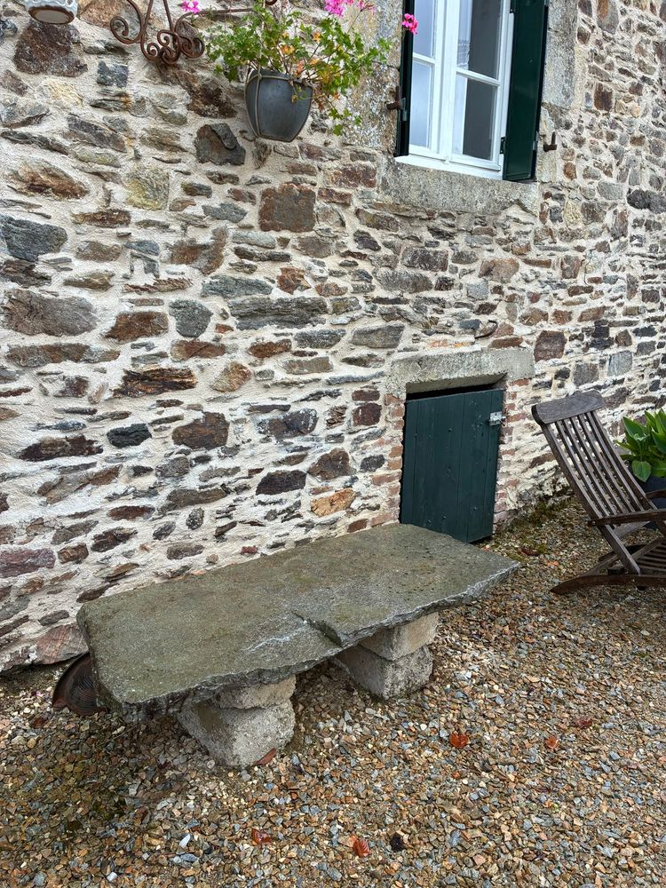 Voor een oude Franse gevoegde muur staat een bankje dat is opgebouwd uit een viertal betonblokken met daarop een grote natuursteen. Er boven hangt een geranium, daarnaast een raam met groene luiken. Daaronder een groen kelderdeurtje met daarvoor nog een stukje van een deckchair. Op de voorgrond grind. Alles is nat van de regen.