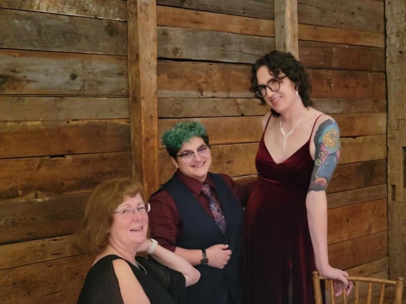 Three people smiling at the camera, an older woman in a black dress seated, a green haired butch in a red shirt and navy vest, and a beautiful tall femme in a red velvet dress