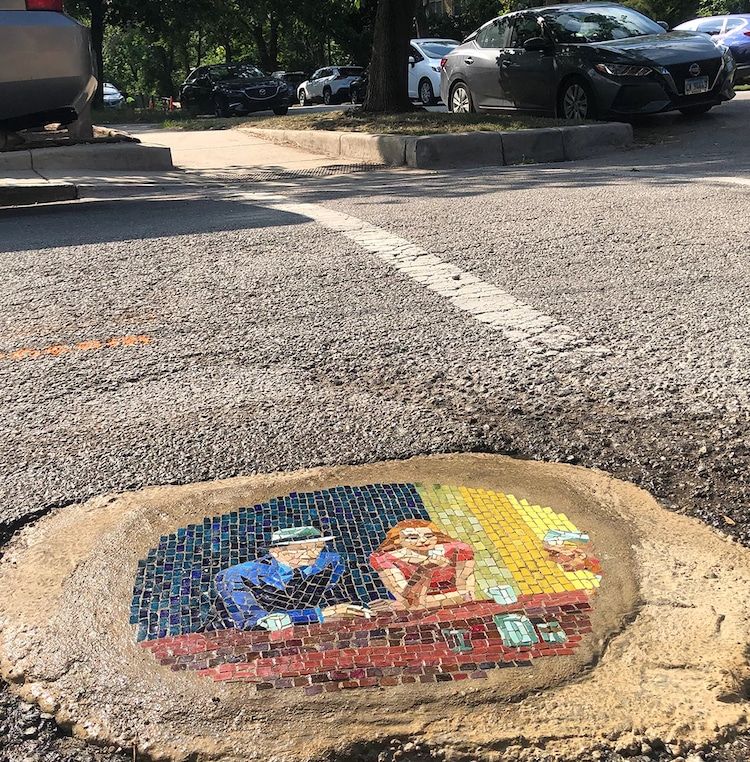The image shows a mosaic artwork embedded in a pothole on a street. The mosaic depicts two figures sitting at a table, with a blue background behind the man and a yellow background behind the woman. The man is wearing a blue shirt and a hat, while the woman is wearing a red dress. The table is represented by red tiles, and there are various objects on it, including a green bottle and a blue cup. The surrounding area includes a parking lot with several cars, including a black Nissan and a white car, and a sidewalk with trees in the background. The street has a white line marking the edge of the parking lot. The mosaic is surrounded by a rough, beige-colored border, indicating the pothole's edge.

Provided by @altbot, generated privately and locally using Ovis2-8B