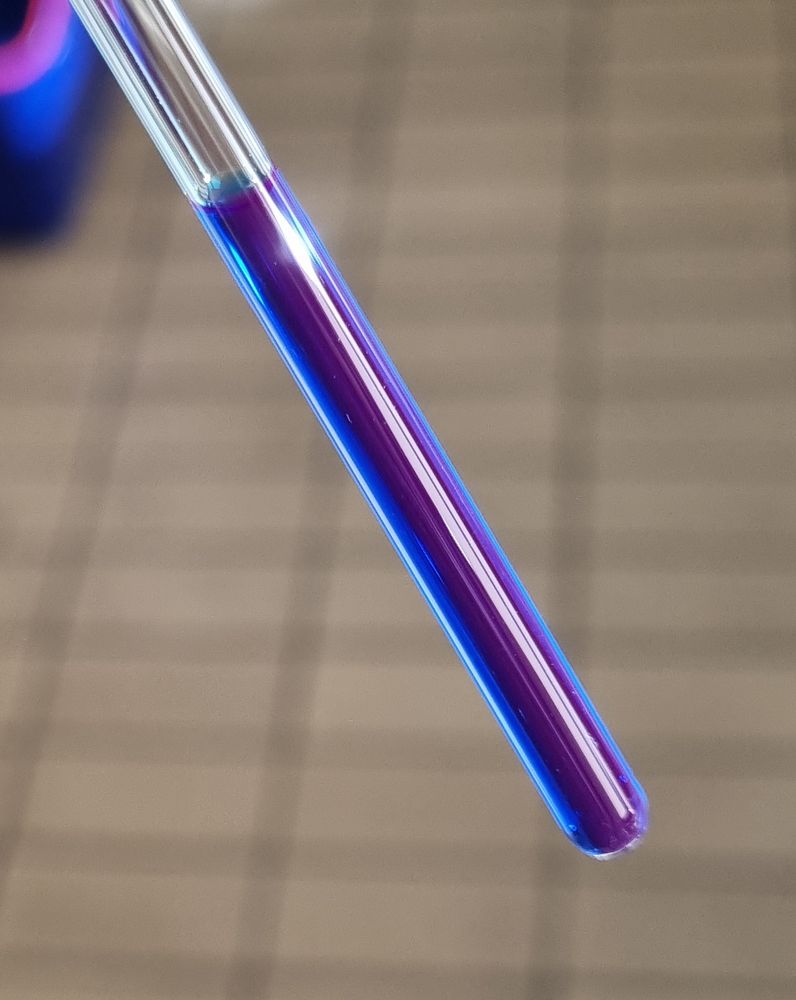 A closeup image of a small cylindrical glass tube used for NMR spectrocopy filled with a blue dye solution that shines a faint pinkish light.