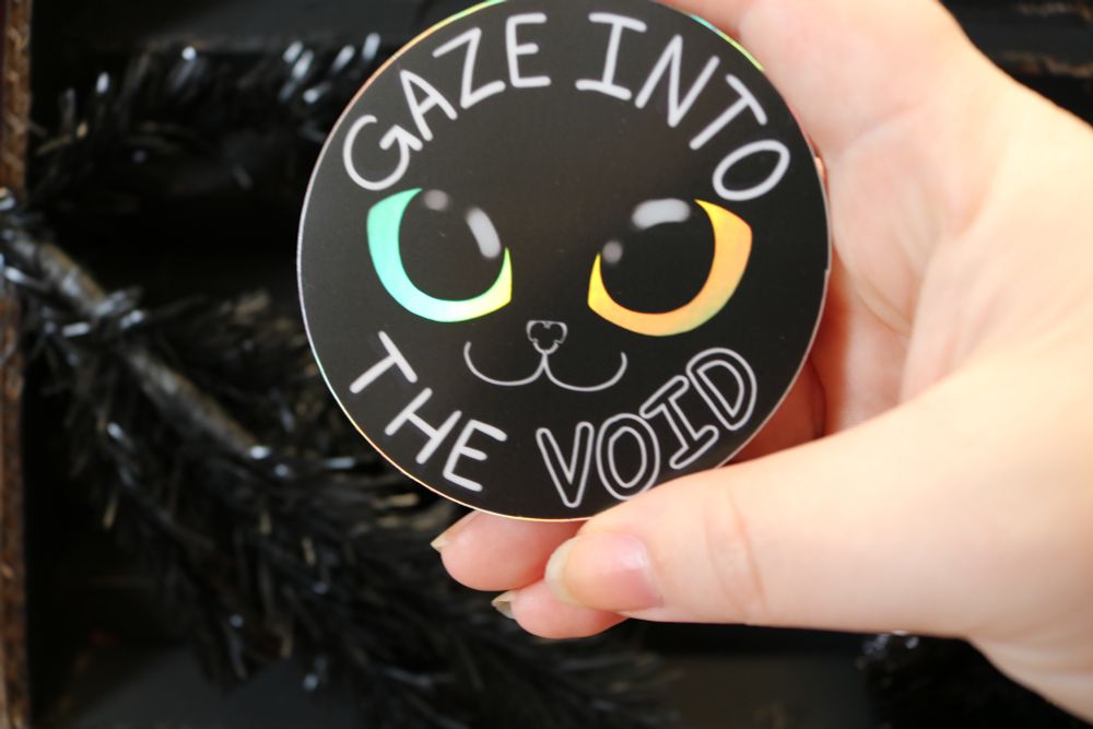 A photo of a 3 inch circular sticker with a cat face on it. The majority of the sticker is matte black, but the cat's eyes are a reflective holo. The text reads "Gaze into the VOID".