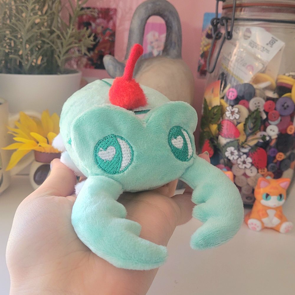 A small light green beetle plush held in the hand of the artist. It is inspired by the colors of melon cream soda and has big green eyes with white heart-shaped sparkles. A red pompom cherry is sewn onto its back. 