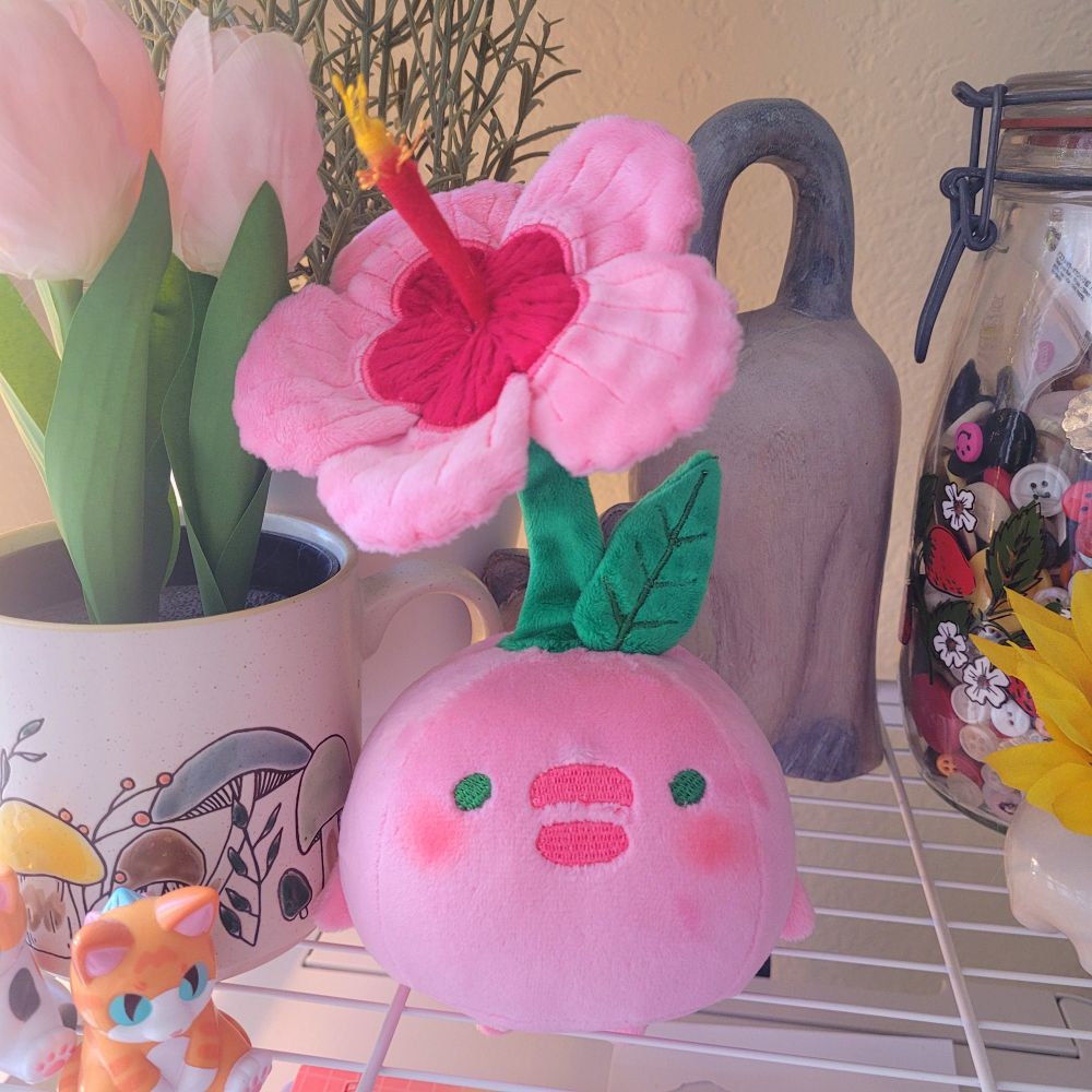 A picture of a round pink plush with a pink hibiscus flower on a green stem. Its cheeks have bright pink blush.