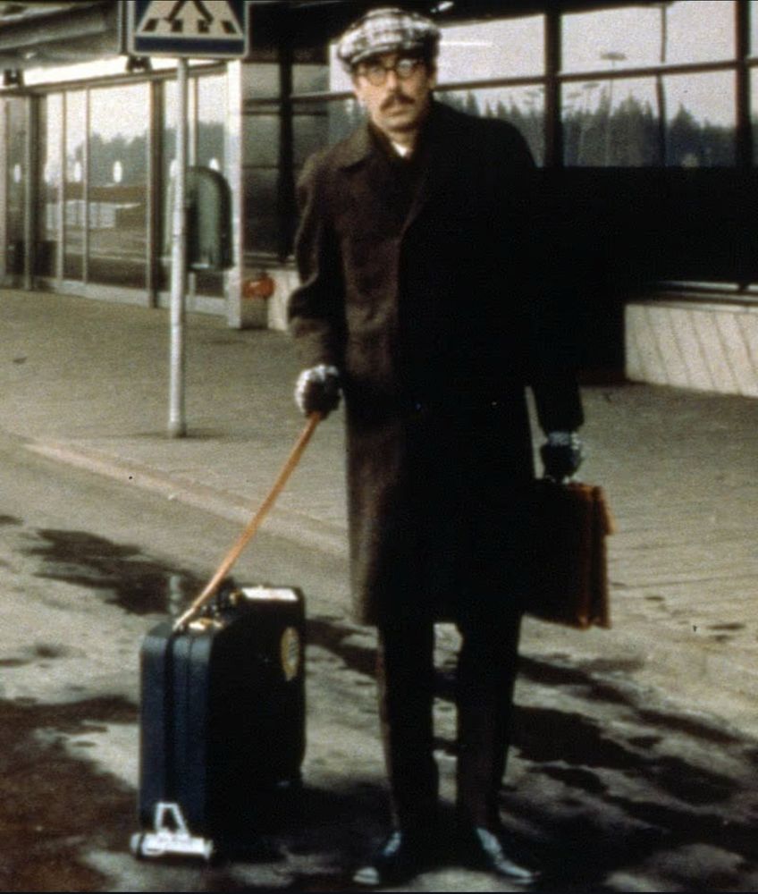 Stig Helmer med packad resväska, lång rock och gubbkeps 