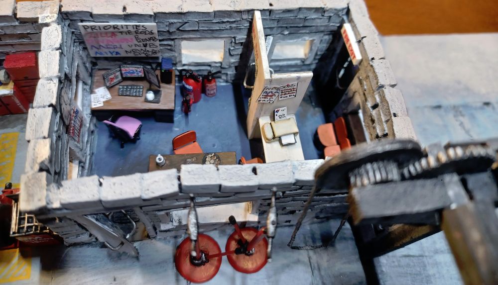 If you open this image in a new window you can see that Laura has a weapons deisgn program open on her left screen, a CAD project with a car in the centre and something that looks suspiciously like Discord open on the right. You can also see the slightly-yellowed old all-in one print/fax/copy/phone thing on the reception desk that screams like hell when it's running but Laura refuses to replace it because she knows how to refill the toner cartridges herself, and in the foreground on the right of the image you can see the hand-cut gears that "operate" the roller shutter.

The roller shutter cable is a cotton cord, again stiffened with cuperglue to pull it into a more realistic-looking shape.