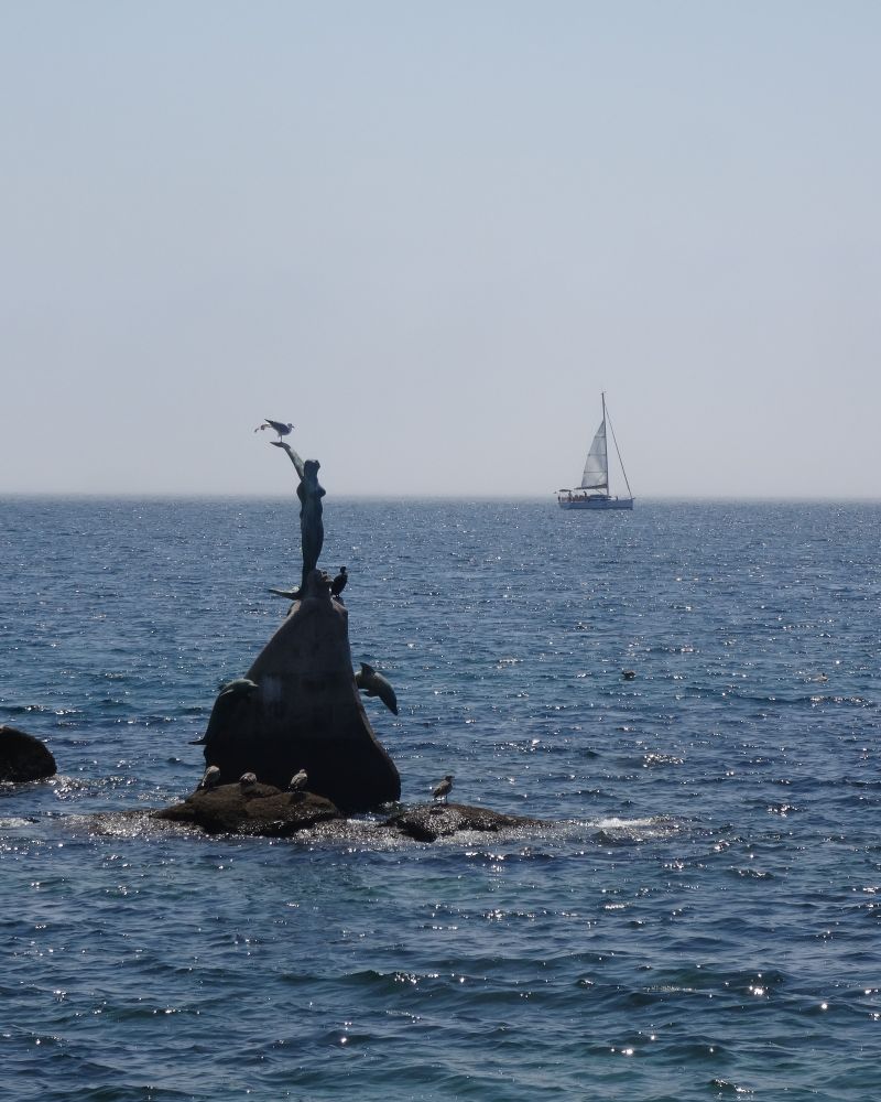 Escultura de una sirena sobre una piedra en el mar en primer plano con un velero sobre la línea del horizonte en segundo plano.