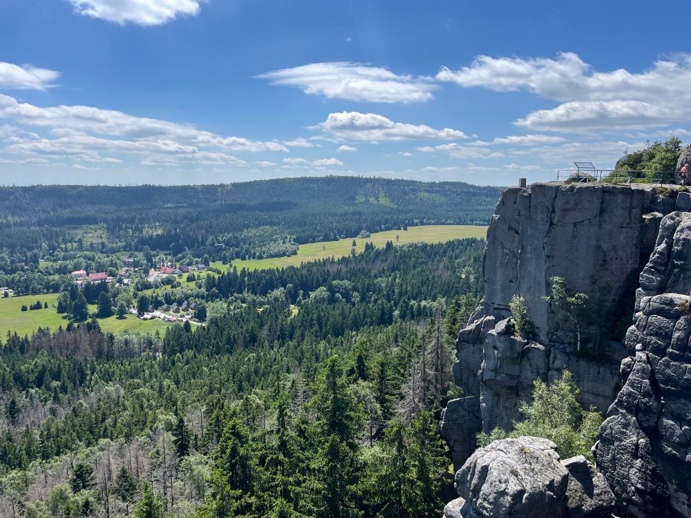 Rocky cliffs and a valley below. It’s a warm and sunny day, and the valley looks full of life. There are a few scattered buildings in the valley but other than those, it’s mostly forest and meadow
