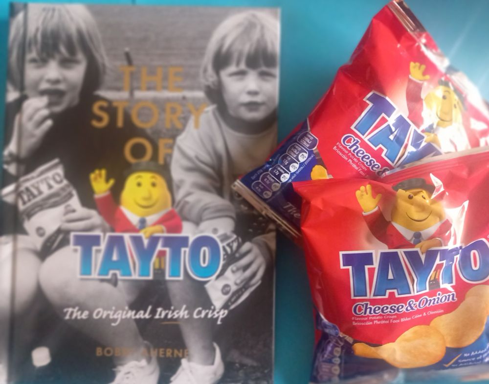 cover image of The Story of Tayto  by Bobby Aherne, as well as two packs of Cheese & Onion Tayto. 