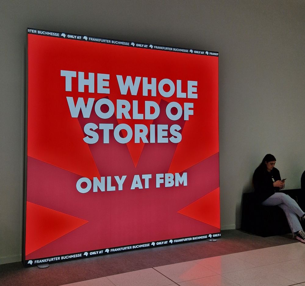 Cartel luminoso donde se lee 'The whole world of stories only at frankfurt book messe' Al lado hay una chica consultando su móvil en uno de los bancos del pasillo. 