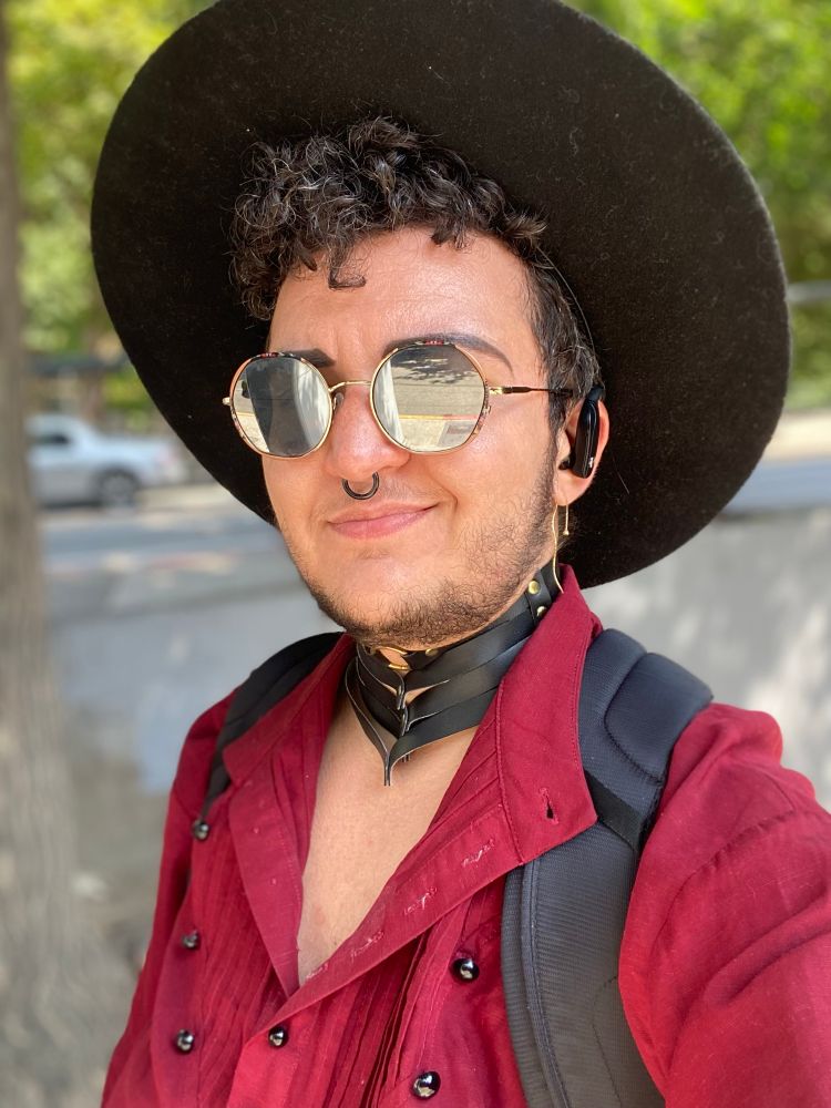 selfie of myself. it’s a lovely sunny day out and I’m wearing a loosely-buttoned, fruity red blouse with a gold multi-strip leather choker, round reflective sunglasses, and my favorite black wide-brim stetson hat