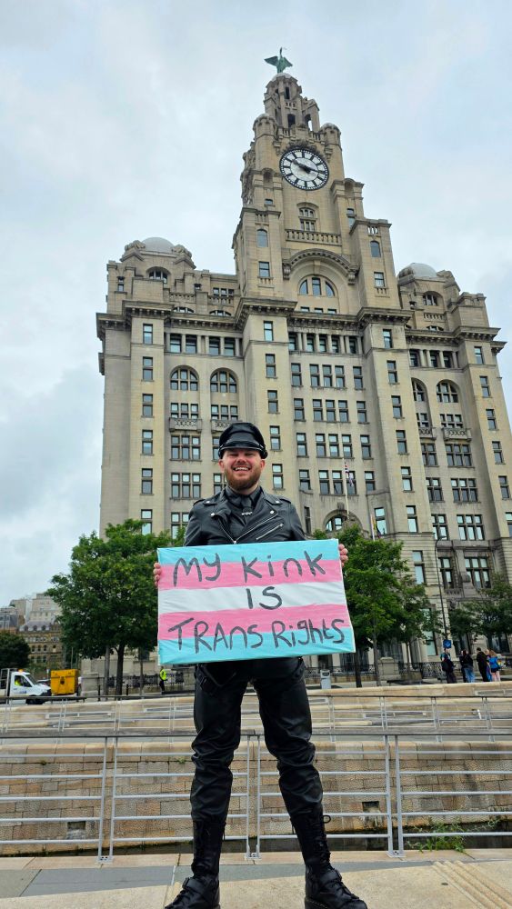 A leather man with a sign that says "My kink is trans rights". He is Standing in front of the Royal Liver Building.