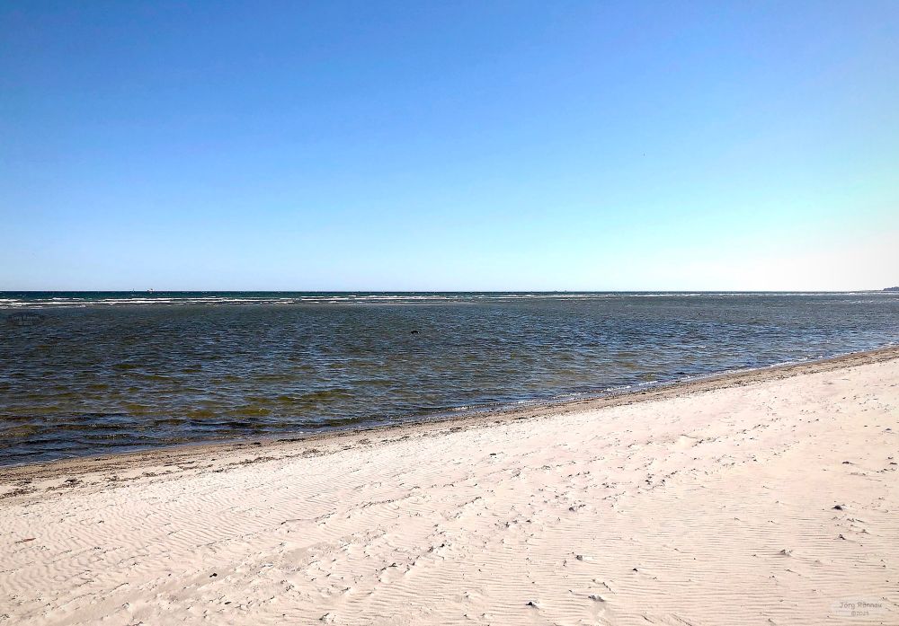 Das Bild zeigt einen Strand bei strahlendem Sonnenschein. Der Himmel ist klar und blau. Das Meer ist ruhig und leicht gewellt. Der Sand ist hell und weist Spuren von Wellen auf.