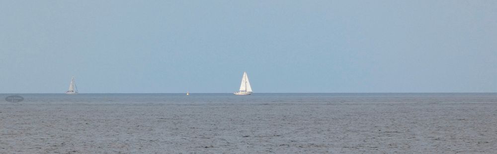 Das Foto zeigt ein weites Panorama der Ostsee. Auf dem ruhigen Meer fahren zwei Segelboot und eine gelbe Boje ist am Horizont zu sehen. Der Himmel ist blau. 