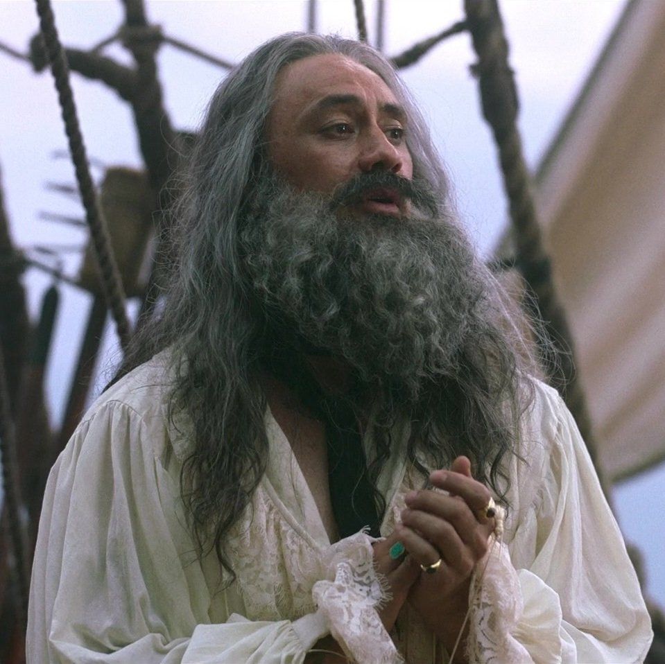 A picture from ofmd season 1 episode 4 of ed standing on the deck in stede's clothes with his hands clasped together 