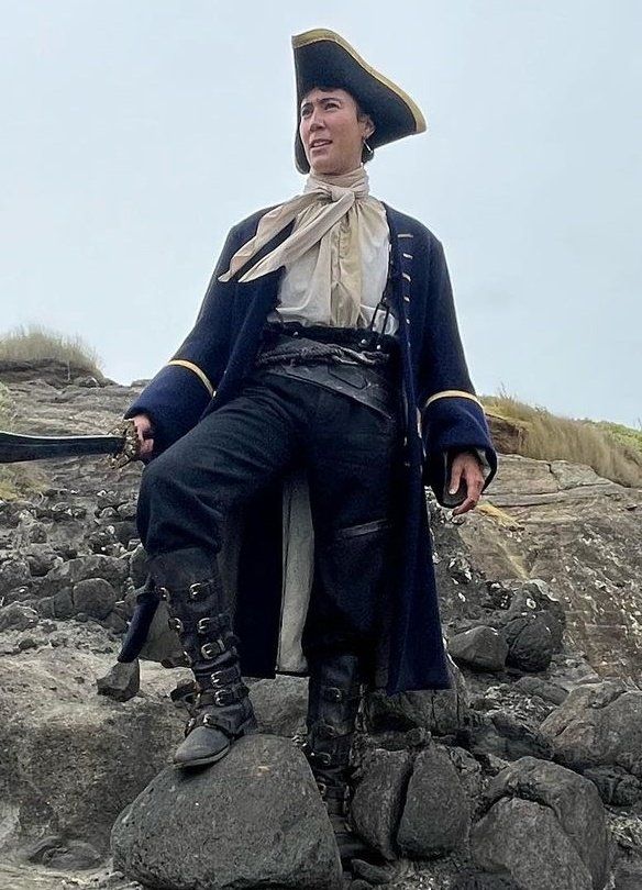 A behind the scenes picture from ofmd season 2 episode 8 of jim standing in a wall of rocks and holding a sword wearing the british navy uniform