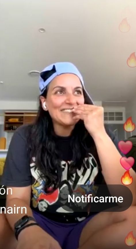 A screenshot from a wee john wednesday live on kristiannairn's insta of madeleine sami smiling big and looking away with a hand on her mouth, while wearing a backwards light blue cap, a black shirt and blue shorts 