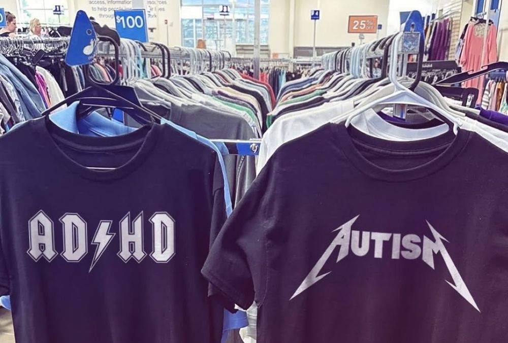 A pictures of two graphic black shirts next to each other in the rack, the text in both is rock band shirt style, the first one says "ADHD" and the second one "AUTISM"